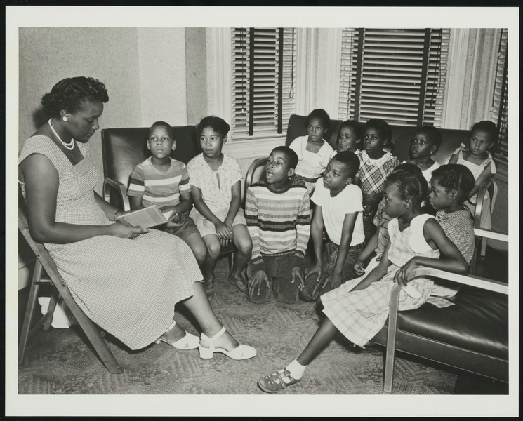 Image of an adult Black female reading to a group of Black children seated in chairs and on the floor, gathered around her. 
