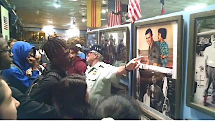 Vietnam veteran wearing hat points to a painting on a display wall, among many, watched by a crowd of students