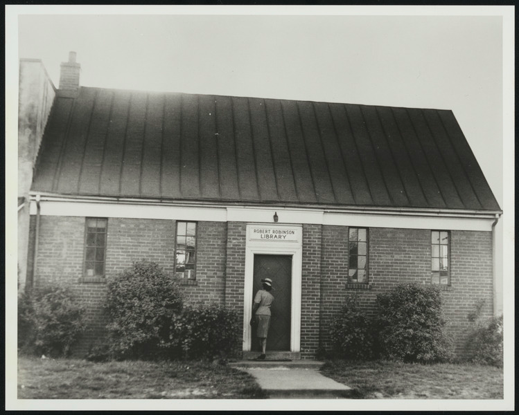 Image of a woman in a skirt and jacket and hat,  entering a library building.