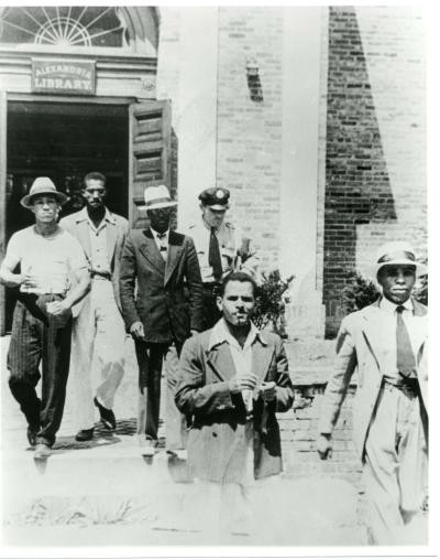 Image of a group of men emerging from a building with police officer. Several men in suits, some wearing hats.