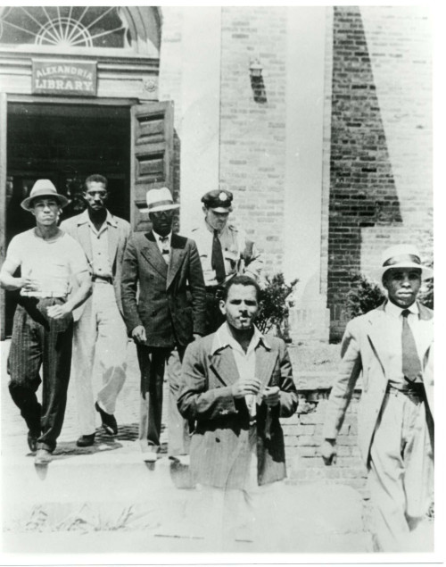 Image of a group of men emerging from a building with police officer. Several men in suits, some wearing hats.